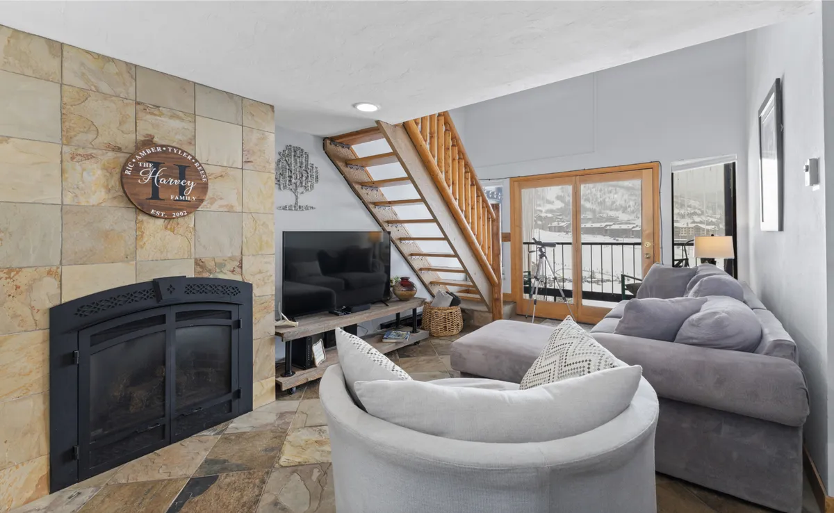 This inviting living room features a plush sofa positioned near a fireplace, with a television and a staircase in the background. Large windows fill the space with natural light and frame stunning views of the surrounding mountains, while soft, neutral tones create a welcoming and relaxing atmosphere.