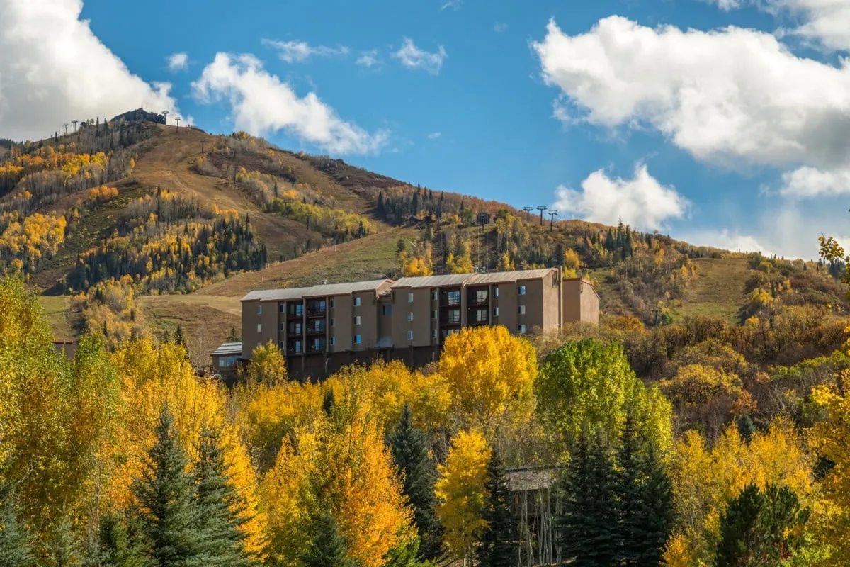 The image shows a charming condo building perched on a hillside, surrounded by vibrant autumn foliage and set against the dramatic backdrop of Steamboat Springs' majestic mountains. The golden hues of fall, panoramic mountain views, and natural beauty immediately catch the viewer's eye, creating a sense of tranquility and adventure.