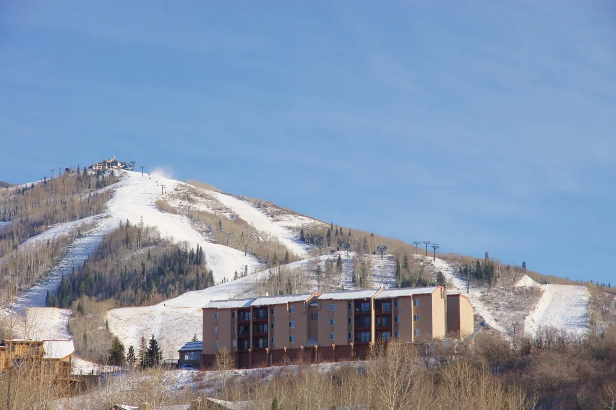 An exterior shot featuring a residential building with the majestic mountain and ski slopes rising behind it. The scene captures the proximity of accommodations to the ski area and highlights both the adventure of the slopes and the comfort of nearby lodging.