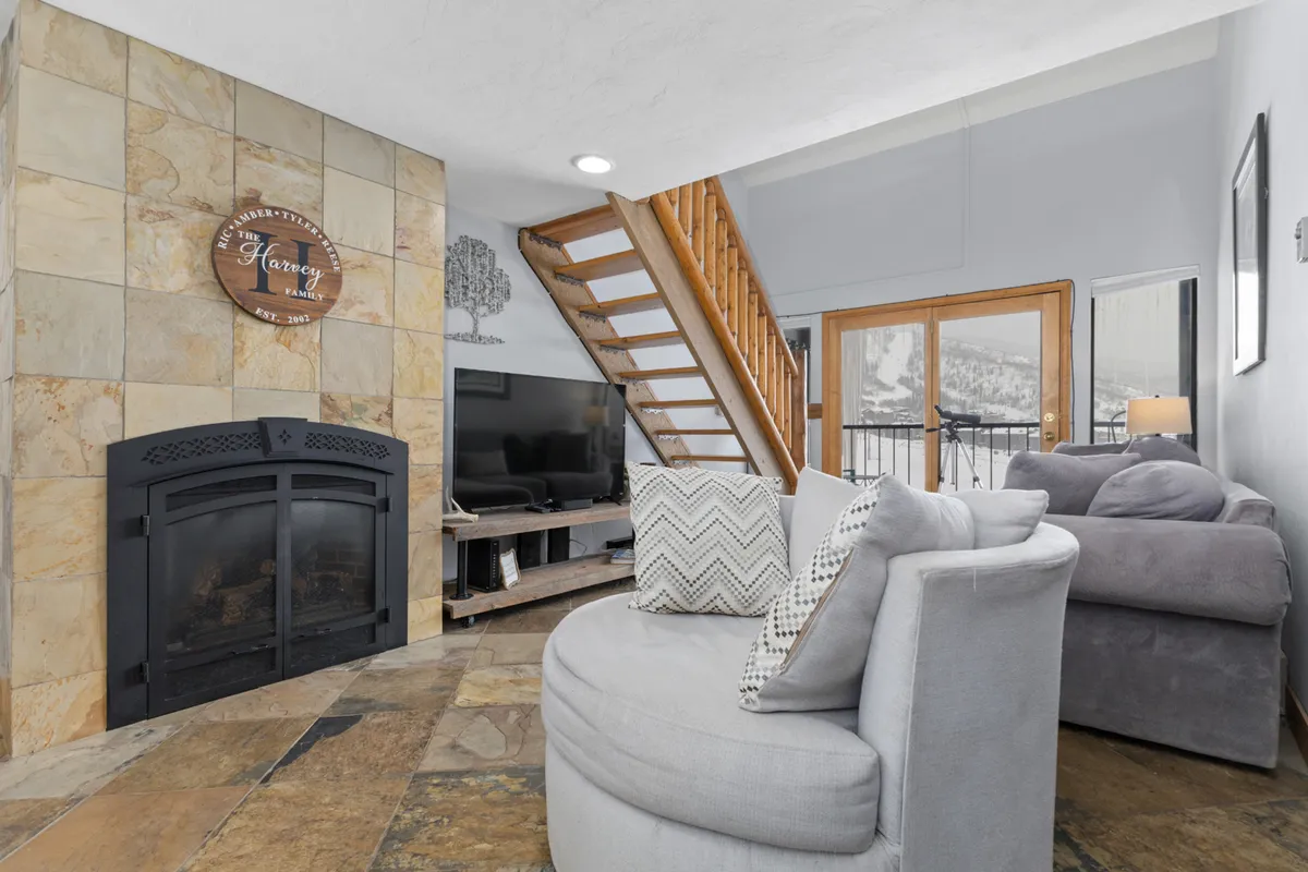 A warm, comfortable living area featuring a fireplace, staircase, and a seating area arranged to take in both conversation and views through large windows. The space is designed for socializing, unwinding after outdoor adventures, and gathering with family or friends.