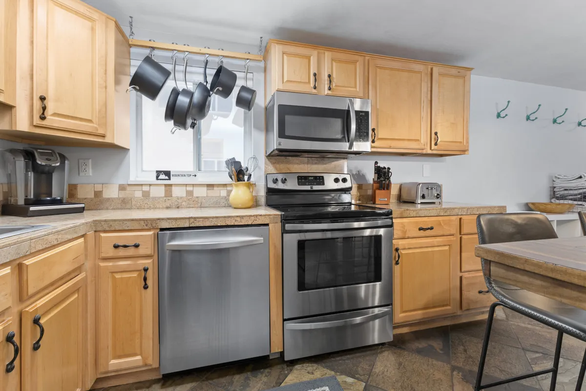This image presents a welcoming kitchen scene with stainless steel appliances, warm wooden cabinets, hanging pots, and a dining table in the foreground. The space looks bright and functional, ideal for preparing family meals, casual dining, or hosting gatherings.