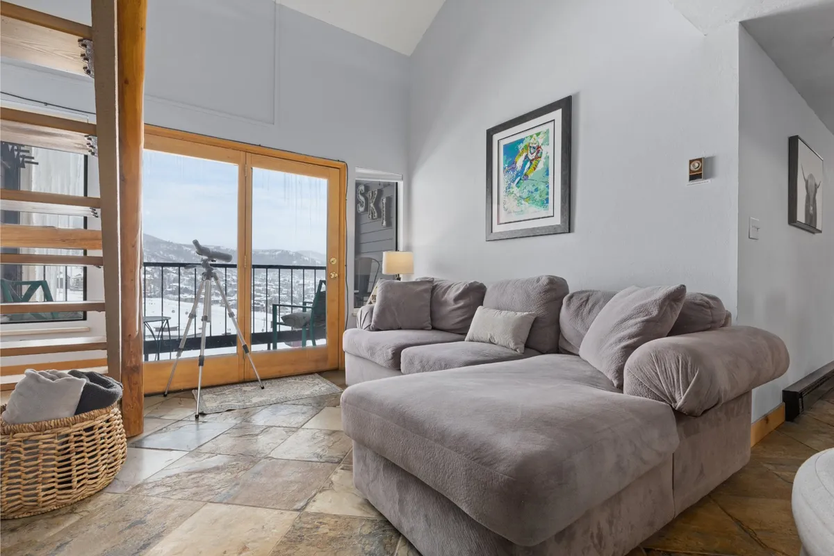 A cozy living space with a large gray sectional sofa faces double glass doors that open to a private balcony, offering a glimpse of scenic outdoor views. The setup encourages both relaxation and easy access to the balcony for enjoying the panoramic Yampa Valley and ski slopes.