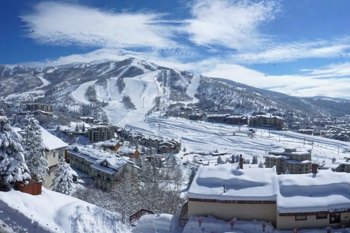A wide, breathtaking view of snow-covered mountain slopes dotted with winding ski runs, pine trees, and residential buildings, all beneath a winter sky. The vantage evokes the thrill and beauty of Steamboat’s ski area in prime winter conditions.