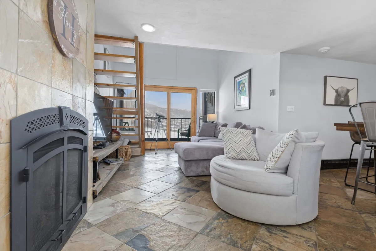 A spacious living room featuring a curved sofa by a central fireplace, a striking spiral staircase, and expansive windows that let in abundant natural light and offer views of the outdoors. The seating area feels inviting and designed for relaxation or social gatherings, perfect for returning from a day on the slopes or enjoying time indoors during any season.