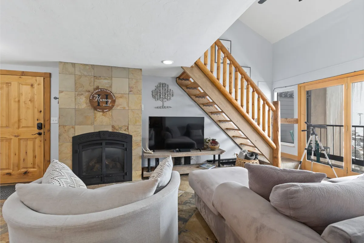 A cozy living area featuring a fireplace, comfortable sofa, staircase, and television. Natural light suggests a nearby balcony or window, hinting at inviting views and a space for relaxation after work or adventures.