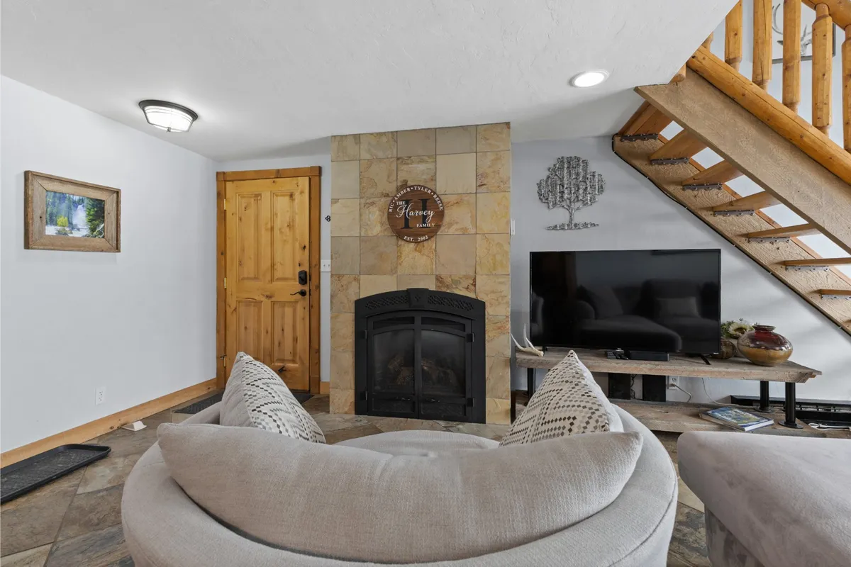 A cozy circular sofa is arranged around a warm fireplace, with a television and decorative wall art creating a welcoming focal point. A wooden staircase adds visual interest and hints at a multi-level layout, while the space invites groups to gather, relax, or enjoy movie nights together.