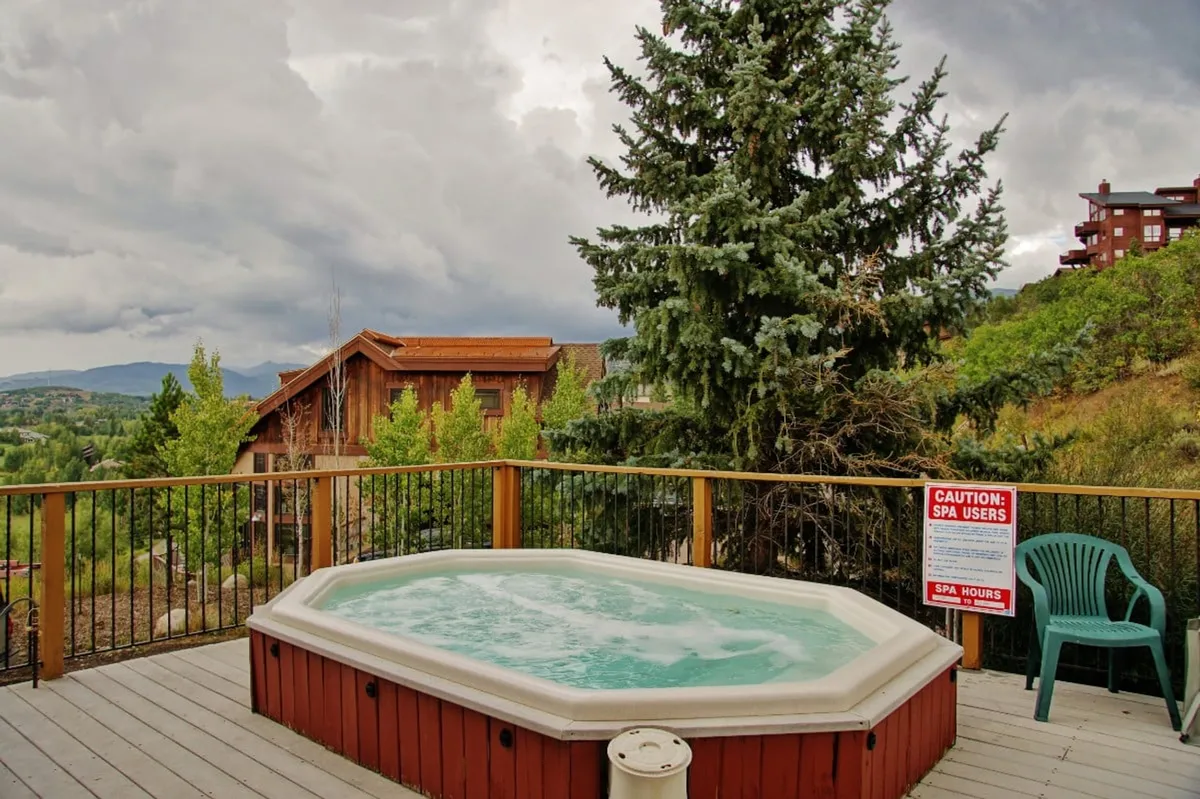 A tranquil exterior hot tub sits on a wooden deck surrounded by lush greenery and mountains, with a few guests unwinding in the warm water and others enjoying coffee. The mood is peaceful yet social, perfect for both relaxation and casual gatherings.