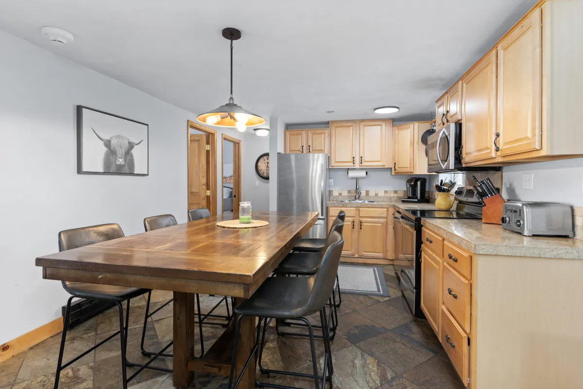A spacious wooden dining table sits at the heart of the room, complemented by kitchen appliances and warm-toned artwork. The area is inviting, perfect for family meals, casual get-togethers, or working on a laptop.