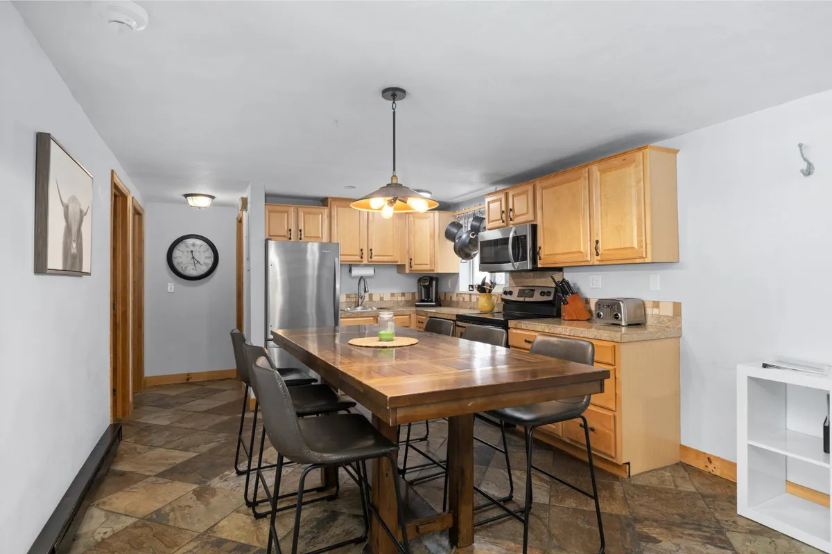 A spacious dining area with a large wooden table is seamlessly connected to a modern kitchen, equipped with stainless steel appliances and illuminated by a warm overhead light fixture. The setting is ideal for shared meals, celebrations, or casual gatherings with friends and family.
